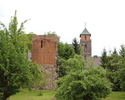 Zdjęcie przedstawia basztę prochową w Baniach. Na pierwszym planie widać basztę wśród zieleni. W tle widoczna jest wieża kościoła.                                                                      