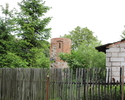 Zdjęcie przedstawia basztę prochową w Baniach. Na pierwszym planie widoczny jest płot ogradzający teren, na którym mieści się baszta,  po prawej stronie widoczny jest fragment sąsiednich zabudowań.   