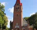 Zdjęcie przedstawia stronę wejścia kościoła wykonanego z czerwonej cegły. Kościół ogrodzony wysokim płotem.                                                                                             
