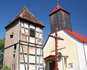 Na zdjęciu widać kościół, osobną szachulcową dzwonnicę na podmurówce z kamienia polnego oraz drewniany krzyż witający odwiedzających.                                                                   