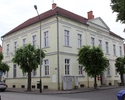 Zdjęcie przedstawia gmach poczty w Trzcińsku-Zdroju. Na pierwszym planie widać boczną i frontową elewację budynku. Zabytek częściowo jest przysłonięty przez drzewa.                                    