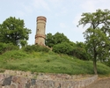 Zdjęcie przedstawia wieżę widokową w Cedyni. Na pierwszym planie widać mur, który otacza wzgórze, w tle wieża w otoczeniu zieleni.                                                                      