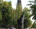 Zdjęcie przedstawia teren starego miasta. Na pierwszym planie widać pomnik, który znajduje się na placu przed ratuszem, w tle, nad drzewami wieża kościoła Mariackiego.                                 