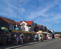 Zdjęcie przedstawia główną drogę w miejscowości Dąbki wraz z zabudowaniami i przyulicznymi sklepikami.                                                                                                  