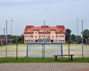 Zdjęcie przedstawia boisko wielofunkcyjne z bramkami do piłki nożnej. W tle widać pomalowany na pomarańczowo blok mieszkalny.                                                                           