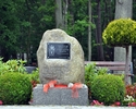Zdjęcie przedstawia głaz na granitowej podstawie, z kamienna, grawerowaną tablicą pamiątkową, otoczony zielenią, uchwycony w ujęciu frontowym.                                                          