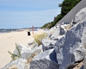 Zdjęcie przedstawia zmodernizowaną, betonową, podsypaną dużymi kamieniami linie brzegową, piaszczystą plażę oraz morze                                                                                  