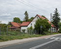 Zdjęcie przedstawia dom nr 6 w Starych Łysogórkach. Na pierwszym planie widoczna jest główna droga przebiegająca przez wieś, dalej otaczająca dom zieleń i boczna elewacja zabytku.                     