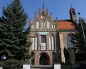 Zdjęcie przedstawia kościół od strony wejściowej, który został wykonany z jasnej cegły. Dookoła parafii znajduje się zieleń.                                                                            