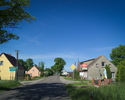 Zdjęcie przedstawia główną drogę we wsi Zakrzewo wraz z zabudowaniami.                                                                                                                                  