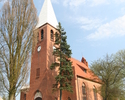 Zdjęcie przedstawia stronę wejścia kościoła zbudowanego z czerwonej cegły, usytuowanego na wzniesieniu. Kościół ogrodzony jest białym murowanym płotem.                                                 