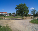 Zdjęcie przedstawia główną drogę we wsi Złakowo wraz zabudowaniami i placem zabaw.                                                                                                                      