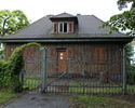 Zdjęcie pokazuje front małego, drewnianego domku ogrodnika, a przed nim ogordzenie, częściowo porośnięte bluszczem.                                                                                     