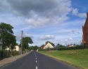 Zdjęcie przedstawia główną drogę we wsi Tychowo wraz z zabudowaniami. Na zdjęciu z prawej strony widoczny jest również fragment kościoła.                                                               