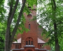 Zdjęcie przedstawia kościół zbudowany z czerwonej cegły uchwycony od strony głównego wejścia, po którego obu stronach rosną wysokie drzewa.                                                             