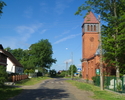 Zdjęcie przedstawia główna drogę we wsi Pałowo wraz z zabudowaniami. Na zdjęciu z prawej strony widoczny jest również kościół i fragment cmentarza.                                                     