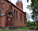 Zdjęcie przedstawia kościół z czerwonej cegły od strony wejścia oraz drewniany krzyż                                                                                                                    