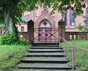zdjęcie przedstawia schody z czerwoną barierką prowadzące do bramy kościoła, otoczonego murem z czerwonej cegły. Za bramą widać główne wrota świątyni.                                                  