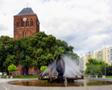Fotografia prxzedstawia fontannę na Placu Jana Pawła II na tle zieleni, kościoła i budynków mieszkalnych.                                                                                               