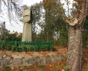 Zdjęcie przedstawia betonowy pomnik w otoczeniu drzew i drewnianym ogrodzeniem na kamiennej podstawie                                                                                                   