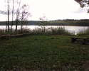 Zdjęcie przedstawia polanę z ogrodzeniem, ławkami i pomostem nad jeziorem                                                                                                                               