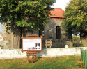 Zdjęcie przedstawia tablicę informacyjną oraz kościół z kamienia w otoczeniu drzew                                                                                                                      