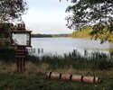 Zdjęcie przedstawia tablicę informacyjną i stojak na rowery na tle jeziora i drzew                                                                                                                      