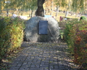 Na zdjęciu widać kamień z tablicą pamiątkowym w otoczeniu zielonego żywopłotu.                                                                                                                          